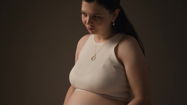 Tilt down studio portrait of young Caucasian pregnant woman embracing her baby bump, representing pregnancy, care and maternal bond