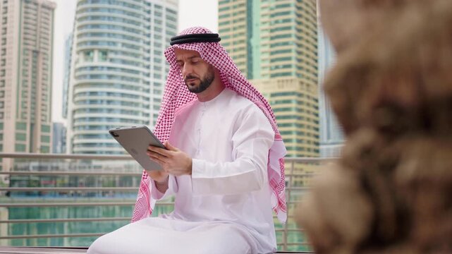Middle Eastern arabic businessman in traditional dress stands in modern city, engaged in planning and investment using a tablet for future success.
