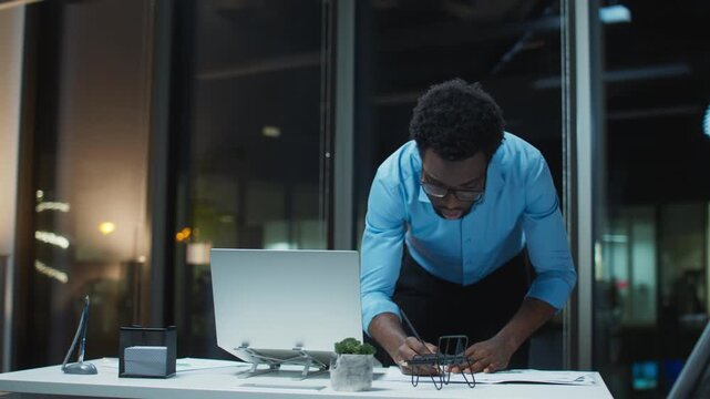 Standing African American male correcting paperwork near laptop. Wearing business shirt and glasses. Reviewing charts on desk. Fixing mistakes under deadline pressure. Late evening inside office.