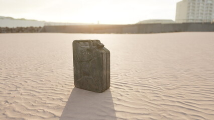 A weathered fuel canister stands alone on the sunlit sandy surface. The golden hour casts long shadows, highlighting its rugged texture and the serene backdrop of a quiet beach area. © icetray