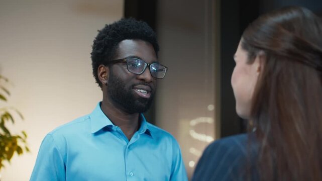 Office colleagues chatting face to face near exit area. Male smiling and speaking. Female listening and reacting warmly. Finishing workday and heading out. Evening inside office building.