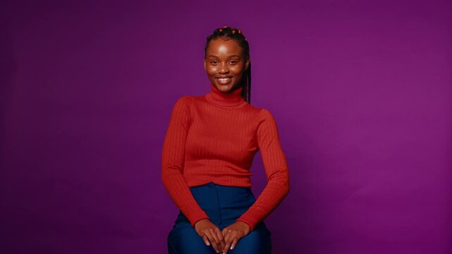 Young black woman smiles and laughs while sitting on a stool against a purple backdrop in a medium, landscape studio shot.