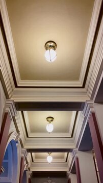Ornate ceiling corridor with warm lights, coffered panels and crown molding, flushmount fixtures casting soft amber glow across arched Taken on mobile device.