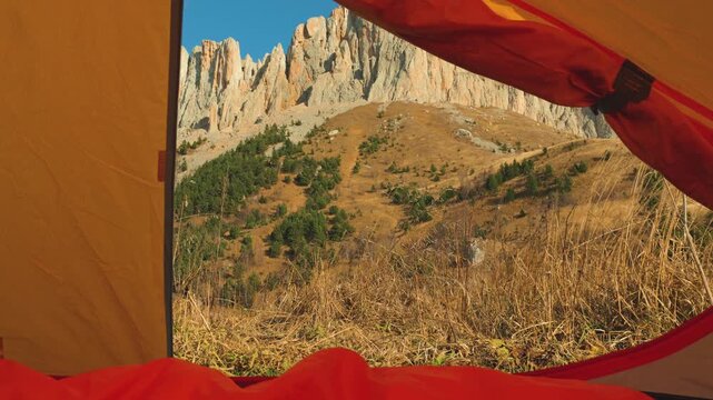 First-person view from inside a tent, with a red sleeping bag in the foreground. The open flap frames a sunny autumn mountain landscape with rocky cliffs. Cozy, adventurous mood