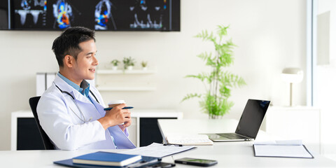 Doctor holding a hot coffee drink cup working in office, using a laptop to access patient records...