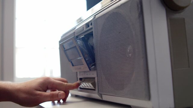 In neon glowing dark studio, hand inserts cassette tape into retro boombox on table, presses play button under warm accent light to activate song. Close-up unrecognizable enthusiast turning on vintage