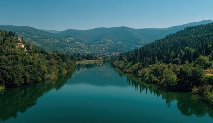 Naklejka premium Vibrant photo of Serene river winding through a dense green forest and rolling hills under a clear blue sky
