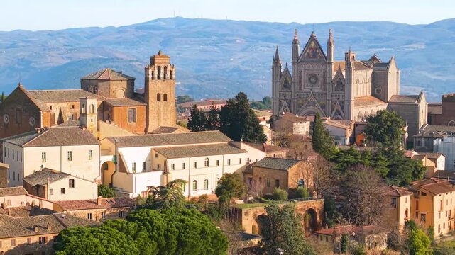 Aerial reveal of Orvieto Duomo facade behind historic church. Drone flight over medieval rooftops and monastery to Orvieto Cathedral in Umbria Italy. 4K 10-bit HDR cinematic morning video.