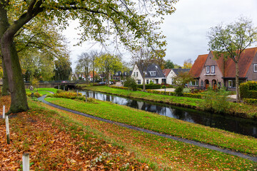 Naklejka premium Hattem town Netherlands canal brick houses autumn foliage scenic idyllic landscape view. Quiet residential neighborhood with trees reflections pathway under cloudy sky peaceful travel atmosphere