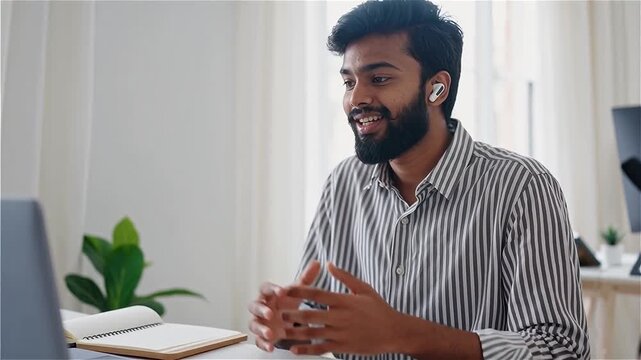 Young Indian Man Working From Home With Wireless Earbuds And Laptop, Engaging In Conversation, Modern Lifestyle, Remote Work

