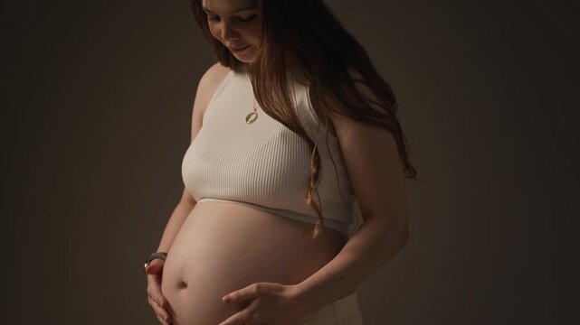 Tilt up medium portrait of young Caucasian expecting mother stroking her baby bump in soft studio lighting, expressing tenderness, anticipation and prenatal care