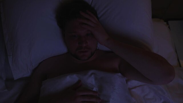 Worried man lying awake in bed at night, suffering from insomnia and anxiety, unable to sleep due to stress, touching his forehead and feeling restless in the dark bedroom, unable to sleep