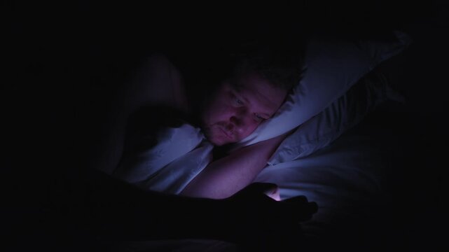 Sleepless man suffering from insomnia waking up in the middle of the night to check his smartphone, addicted to social media and suffering from nomophobia and sleep disorder