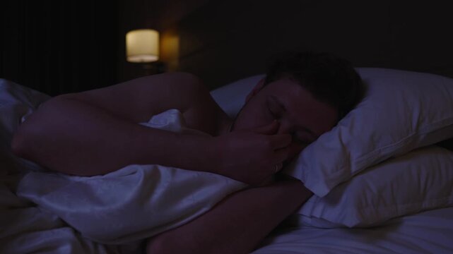 Annoyed man turning off the alarm on his smartphone in the morning, getting ready to wake up and start a new day, but wanting to continue sleeping in his comfortable bed under the warm covers