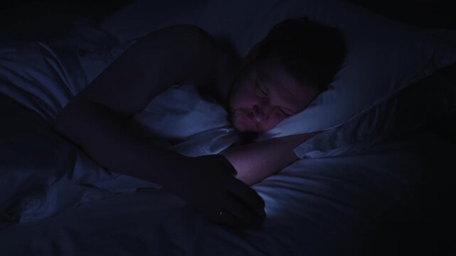 Sleepless man with phone addiction scrolling through social media feeds in dark bedroom at night, struggling with insomnia from the bright screen light and blue light exposure