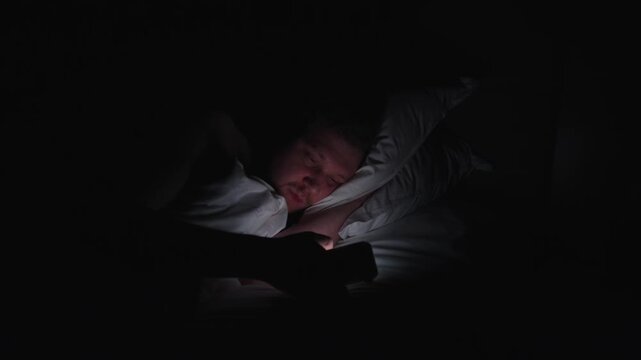 Lonely man lying in dark bed at night, illuminated by the bright screen of his smartphone as he scrolls, suffering from insomnia and phone addiction while doomscrolling social media