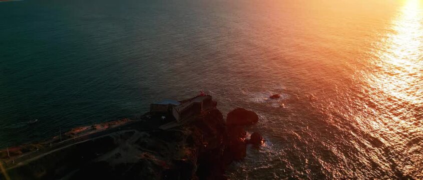 Breathtaking aerial drone footage of the historic Forte de S&atilde;o Miguel Arcanjo lighthouse in Nazar&eacute;, Portugal. Atlantic waves crash against the cliffs during a stunning golden hour sunset.