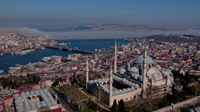 Aerial drone view of S&uuml;leymaniye Mosque in Istanbul, featuring its domes and minarets with the Golden Horn, bridges, boats and historic city skyline in the background.