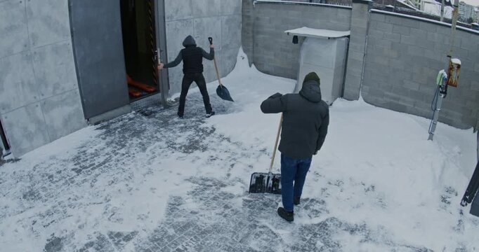 Two Men Shovel Snow on Yard Near Open Garage With Orange Pallet Jack Visible Inside. High Angle CCTV Captures Winter Facility Maintenance Beside Brick Wall, Showing Routine Outdoor Work Property Care.