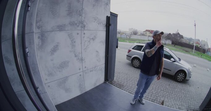 Wide Angle Doorbell View of Courier With Tattoos on Arm, Smoking and Checking Phone While Waiting at Door. Concept of Smart Home Security, Mobile Delivery Tracking, Modern Logistics Delivery Services.
