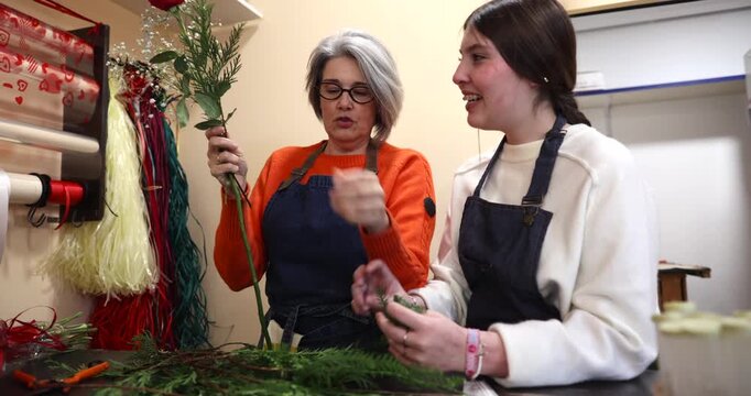 Experienced senior florist teaching a young female apprentice how to create a beautiful floral arrangement, sharing her professional skills and knowledge inside a cozy flower shop workshop