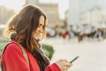 Smiling curly young woman in red sweater looking at smartphone screen on sunny city street.