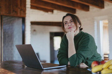 Mature caucasian woman dreaming and smiling while working on laptop at home office