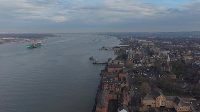 Container ship on river Thames sailing by Gravesend town. Aerial view of large green vessel escorted by pilot boat towards the Docks near London. Gravesend town with historic church and pier.