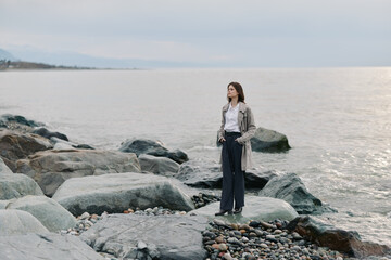 A thoughtful woman stands on rocky shore looking at calm sea horizon. She wears a trench coat and dark pants in overcast daylight. Concept of solitude, reflection, and nature outdoors.