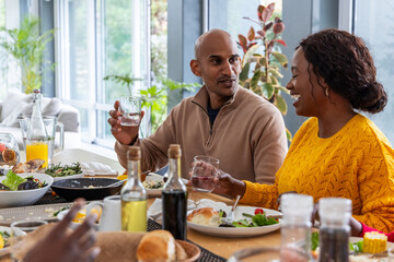 Diverse midlife couple in beige pullover and yellow knit chatting at dining table holding glasses