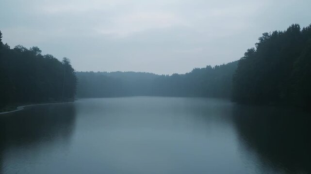 Calm lake reflecting a dense forest on surrounding hills during an early morning with mist lingering over the water, creating a tranquil and atmospheric natural scene