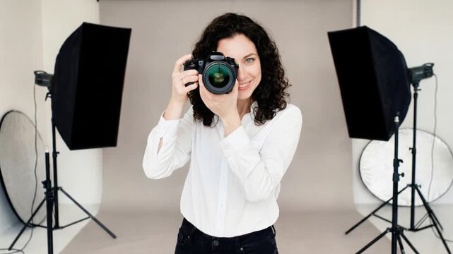 smiling female photographer with curly hair holding professional mirrorless camera in photo studio with lighting equipment