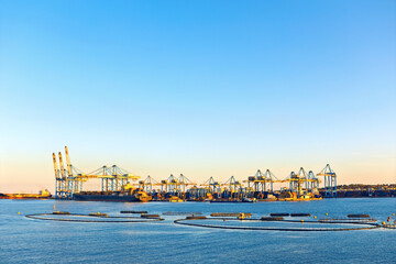 Wide view of a container port with towering cranes beside the sea in Malta. Floating circular containment booms are visible on the calm surface of the water.