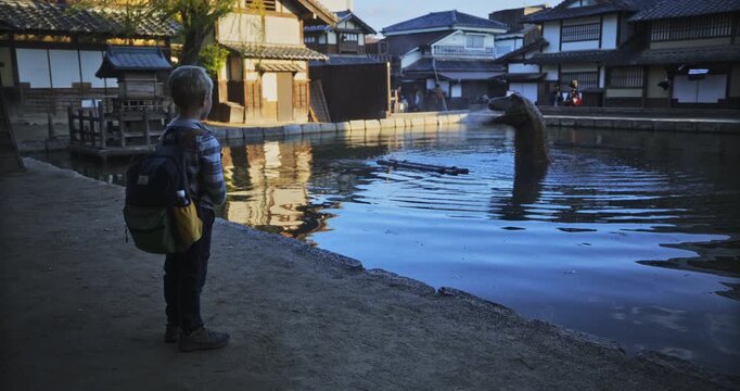 Child with backpack watching dinosaur spewing mist from mouth in pond of old Japanese village