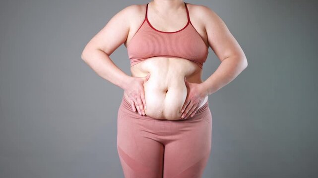 Abdominal massage, tummy tuck and flabby skin on a fat belly, woman doing self-massage with cream on gray studio background