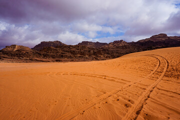 Golden sand dunes with tire tracks and dramatic rocky mountains under cloudy sky in desert landscape at sunset, vast wilderness travel destination scenery. © Duro Mesiar