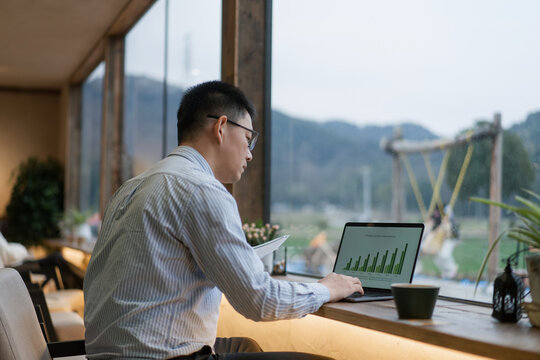 A man wearing glasses analyzes charts on a laptop by the window with mountain view