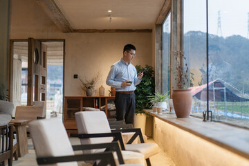 Fototapeta premium A young man wearing glasses stands indoors checking his phone while gazing at mountain scenery outside the window