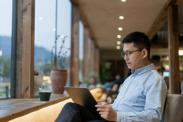 Man wearing glasses using a laptop computer by the window in a cozy indoor setting