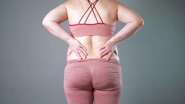 Do-it-yourself back massage, flabby skin and love handles, fat woman doing self-massage on gray studio background