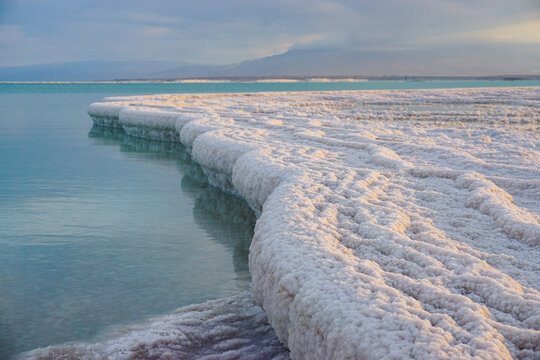 Thick salt crystal formations along calm turquoise water under soft evening light. Natural mineral texture, serene landscape with dramatic sky and distant mountains.