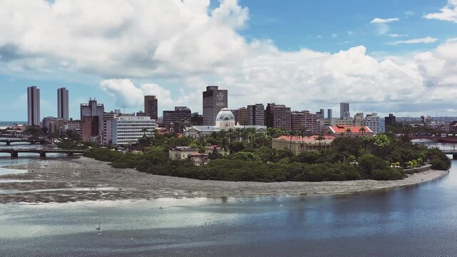 Panoramic view of Recife skyline with modern buildings, bridges, and the Pal&aacute;cio das Flores along the river under a bright blue sky.