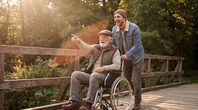 happy caucasian son pushing disabled senior father in wheelchair on bridge. elderly man pointing finger. caregiving and family lifestyle. healthcare, social assistance. banner, website header.
