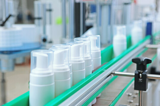 White plastic bottles with dispensers and transparent caps are moving along the conveyor. Manufacture of cosmetics and skin care products. A capping and labeling machine.