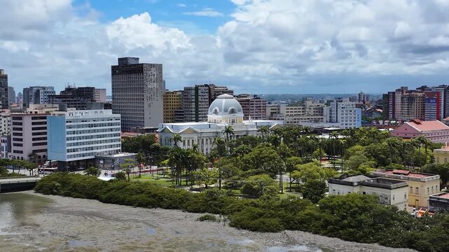 Panoramic view of Recife skyline with modern buildings, bridges, and the Pal&aacute;cio das Flores along the river under a bright blue sky.