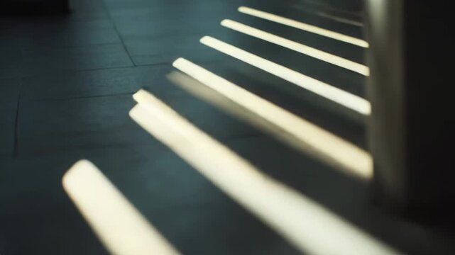 Sunlight streams through a window, casting bright parallel lines of light and contrasting shadow patterns onto a dark, textured floor, creating a minimalist and abstract composition