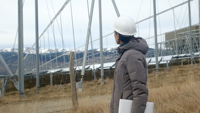 Female engineer wearing a hard hat and winter coat inspecting a concentrated solar power plant with a laptop in hand, working on clean energy generation and industrial infrastructure development