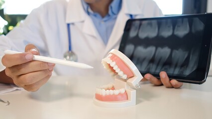 Dentist in scrubs points at a jaw model while showing panoramic dental x-rays on a tablet, explaining oral health, diagnosis and treatment options in a modern clinic