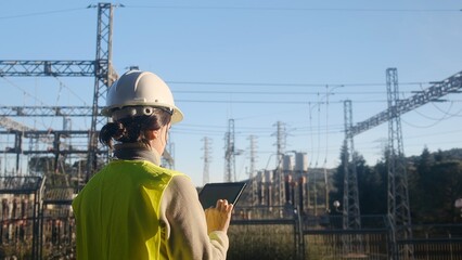 Fototapeta na wymiar Female engineer in safety gear checking infrastructure, using a digital tablet at a power substation, managing and monitoring high voltage electricity grid distribution