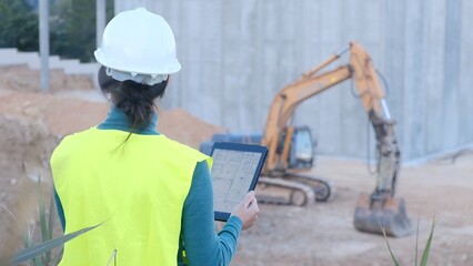 Woman construction professional working on site, reviewing blueprints digitally on portable device, managing project planning and development alongside an excavator © DawDunia